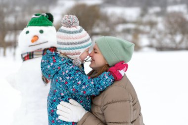 Şapka ve eşarp takan kardan adama karşı anneyi kucaklayan çocuk. Kışın anne ve çocuğun dışarıda portresi.