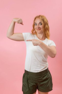 Yeşil gözlü genç kadın kaslarını gösteriyor. Pembe pastel arka planda güzel, güçlü, neşeli, kızıl saçlı bir kız..