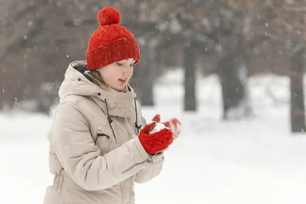 女の子は冬の森でスノーボールをする 赤い帽子の若い女性は雪に覆われた公園の背景で楽しいです — ストック写真