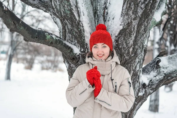 雪に覆われた木の下で美しい若い女性が笑顔でカメラを見つめている 赤いニット帽子とミトンで若い女性 — ストック写真