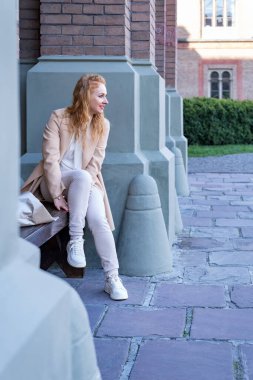 Gülümseyen genç kadın elinde bir buket çiçekle bankta oturur ve birini bekler. Dışarıda yürüyen kızıl saçlı bir kadın. Dikey çerçeve