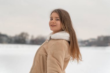 Uzun saçlı gülümseyen kız, rahat bej kuzu derisi ceketiyle kış doğasının tadını çıkarıyor. Sıcak kadın kıyafetleri..