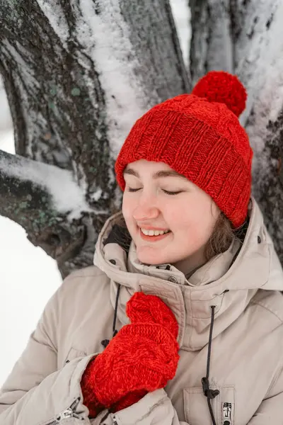 赤い帽子とミトンで笑顔の若い女性は雪に覆われた木の近くに立っています 目を閉じたかわいい思慮深い少女の肖像画 — ストック写真