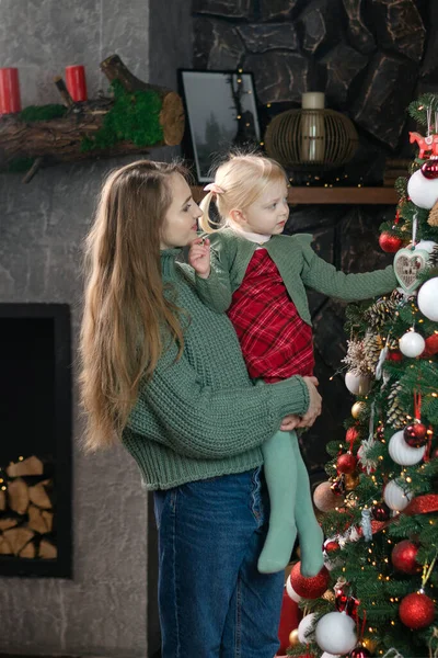 Mom holds little girl near Christmas tree. New Year mood. Mother and daughter decorate Christmas tree at home. Vertical frame
