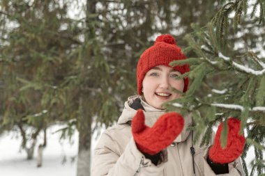 Kış ormanında kırmızı yün şapkalı mutlu bir kız. Kırmızı şapkalı gülümseyen kadın ladin dalına dokunuyor. Karlı orman.