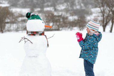 Küçük çocuk kış manzarasında kardan adamın önünde duruyor. Yan görüş. Kış tatilinde çocuk ve kardan adamın portresi