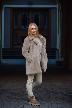Açık renk elbiseli ve kürk mantolu çekici bir genç kadın portresi. Soğuk mevsim, şehirde yürüyüş. Dikey çerçeve.