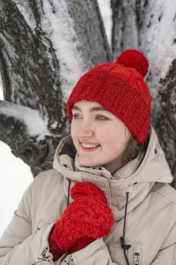 Uzaklara bakan düşünceli genç kızın portresi. Kırmızı örgü şapkalı ve eldivenli genç kadın karla kaplı ağacın yanında duruyor.