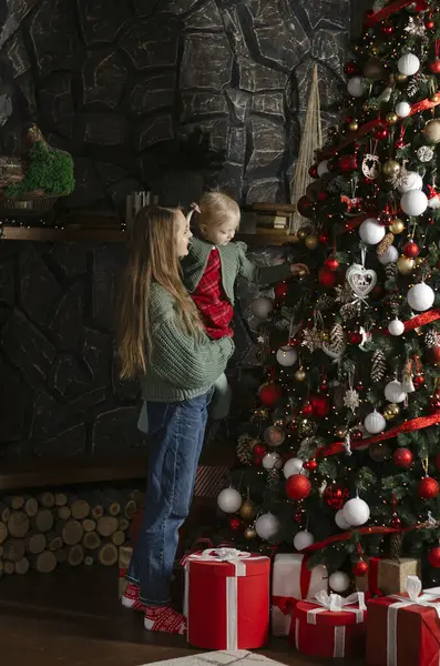 Mom holds little girl on her arms near Christmas tree. Christmas tree at home. New Year mood. Mother and daughter decorate Vertical frame.