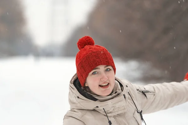 Portrait of smiling girl in red knitted hat with bubo on snow park background. Woman in winter clothes in outside.