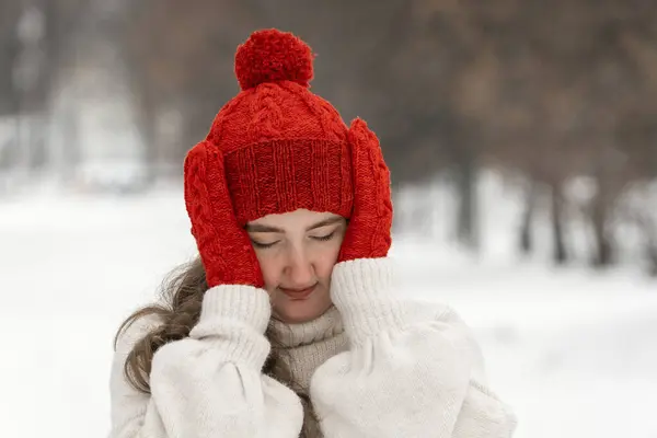 赤い冬の帽子の若い女の子は冬の公園に立っている間彼女の頭を握ります 悲しい若い女性の肖像画 — ストック写真