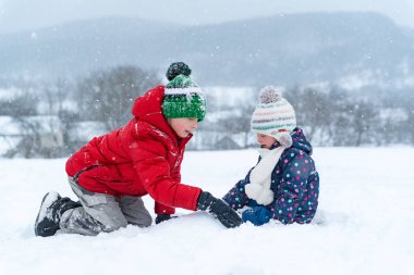 Kırmızı ceketli büyük oğlan ve soğuk bir kış gününde küçük kız kardeş dışarıda kar kalesi heykeli yapıyor. Çocuklar karla oynar, kış tatillerinde.