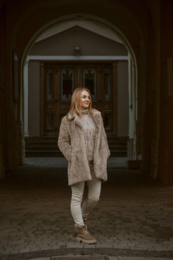 Bir şehir caddesinde, kışlık giysiler ve kürk manto giymiş güzel genç bir kadının tam boy portresi. Soğuk mevsim, şehirde yürüyüş. Dikey çerçeve.