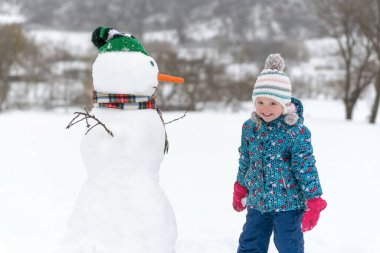 Örgü şapkalı çocuk kışın parkta kardan adam yapıyor. Büyük kardan adam ve mutlu çocuk portresi