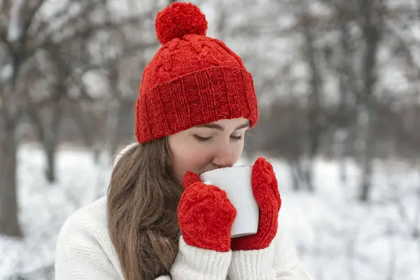 Beautiful young woman drink hot beverage from cup in winter outdoor. Girl in red hat and mittens in snowy park.