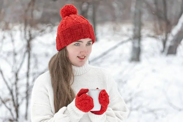 Smiling young woman in red hat and mittens in snowy park. Beautiful girl drink hot beverage from cup in winter outdoor.