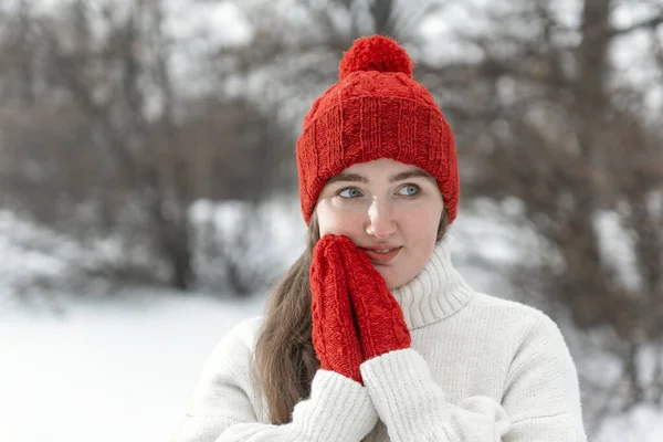 Beautiful woman warms up hands in woolen clothes outdoors, cold weather. Young woman in knitted red hat and mittens, winter park.