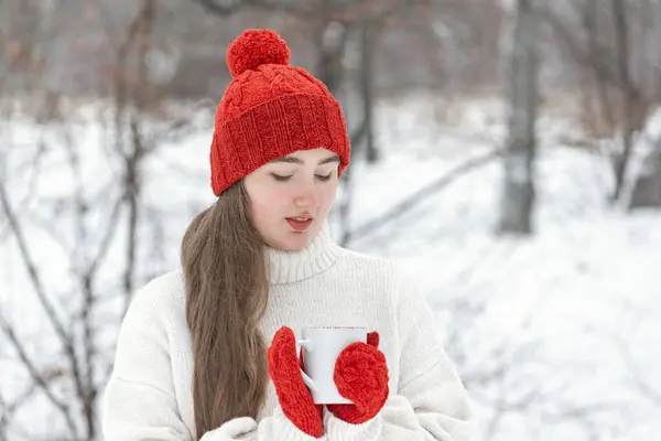 Young woman in red hat and mittens in snowy park. Beautiful girl with cup of beverage in winter outdoor.