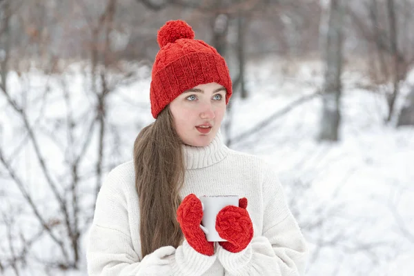 Beautiful girl drink hot beverage from cup in winter outdoor. Smiling young woman in red hat and mittens in snowy park.