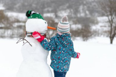 Çocuk geniş bir kardan adama sarılıyor. Çocuklar için kış aktiviteleri. Doğal ışıklandırma rötuş yok