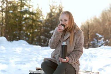 Genç kızıl saçlı kadın kış parkında ya da ormanda elinde elektrikli süpürge şişesiyle çay içiyor. Kış pikniği ve dinlenme