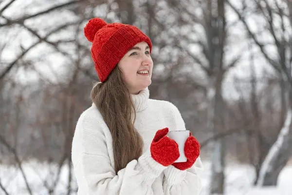 Laughing young woman in red hat and mittens in snowy park. Beautiful happy girl with cup of tea in winter outdoor.