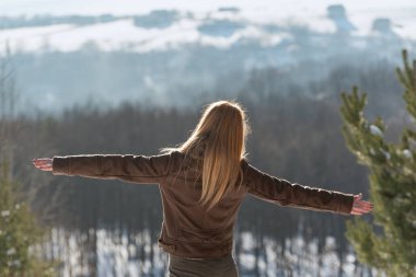 Mutlu genç kadın tepenin üstünde güneşli bir kış gününün tadını çıkarıyor. Arkadan bak. İyi kış havası