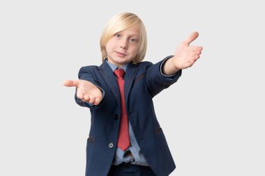 Stüdyo resmi, sarı saçlı, mavi takım elbiseli, kırmızı kravatlı beyaz arka planda elini sıkmak için uzanan beyaz tenli bir çocuk.