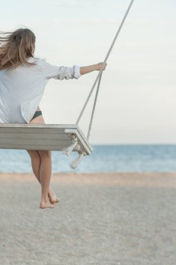 Beyaz tişörtlü kız denizin üstünde sallanıyor. Dikey çerçeve. Arkadan bak. Mavi gökyüzü ufku ve deniz arka planı. Gün batımı yumuşak ışık