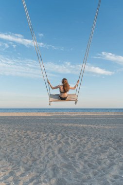 Mayo giymiş genç bir kız mavi gökyüzünde salıncakta sallanıyor, deniz ve plaj arka planında. Dikey çerçeve