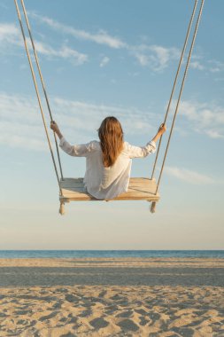 Sarışın, uzun saçlı ve beyaz gömlekli genç bir kadın denizin üstünde salıncakta sallanıyor. Dikey çerçeve. Sarı sihirli günbatımı ışıkları. Arka plan.