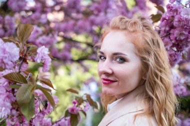 Parkta kırmızı dudaklı şık bir kız. Çiçekli sakura dalları arasında bej ceketli mutlu çekici bir kadının portresi..