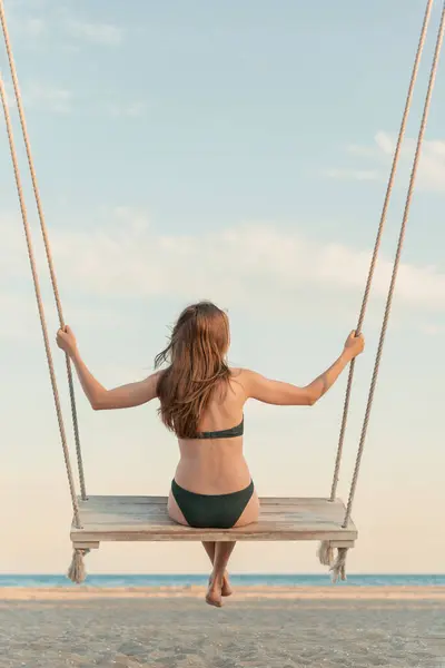 Beautiful young woman with long hair in swimsuit on rope swing high in the sky. Magical light of sunset. Vertical frame. back view