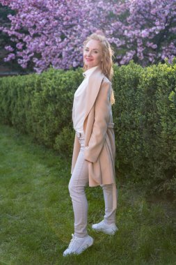 Bahar parkında uzun sarı saçlı, beyaz önlüklü, orta yaşlı, gülümseyen bir kadın. Tam boy portre. Dikey çerçeve