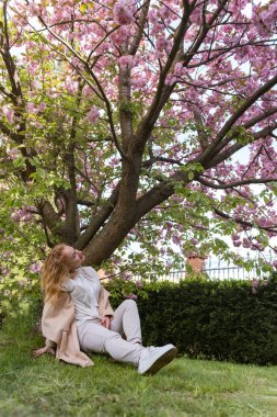 Kız, bahar parkında çiçek açan ağacın altında dinleniyor. Genç kadın kiraz çiçeklerinin altında çimenlerde oturur ve yukarı bakar. Dikey çerçeve