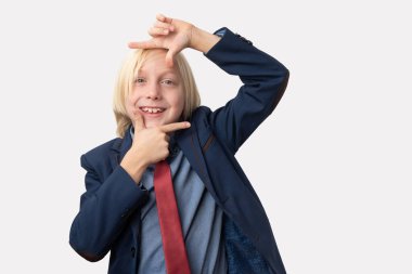 Mavi ceketli ve kırmızı kravatlı şirin bir çocuk beyaz arka planda parmakları olan bir çerçeve yapıyor..