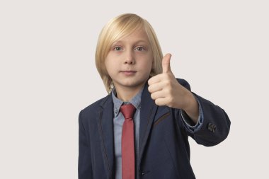 Başparmaklar yukarı. Sarı saçlı, mavi ceketli ve kırmızı kravatlı bir çocuğun portresi, beyaz arka planda izole edilmiş, baş parmak hareketlerini gösteriyor.
