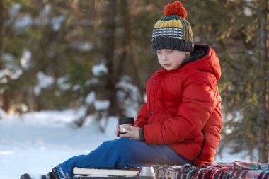 Portrait of boy in warm blown jacket knitted hat with cup of hot tea in winter forest. Picnic in winter in nature clipart