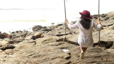 Kırmızı şapkalı sakin kadın deniz kenarındaki kayalık araziye bakan bir ağaç ipinde sallanıyor, yemyeşil ve doğanın sükuneti ile çevrili.