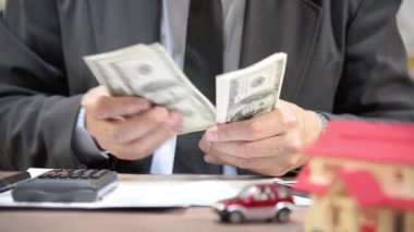 İş adamı hesap makinesi, doküman ve ev ve arabanın küçük oyuncak modelleriyle bir masada dolar sayıyor. Finansal planlama, bütçe veya emlak yatırımını simgeliyor.