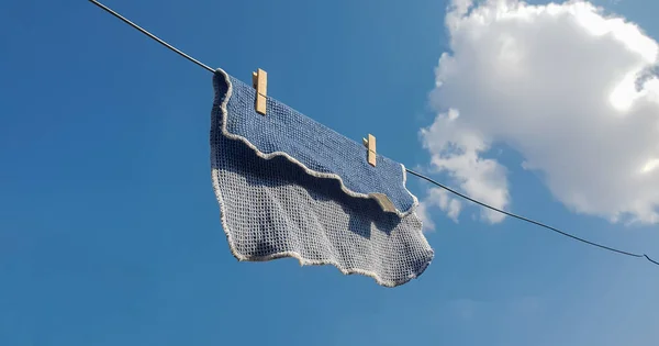 Close up on a dish washing cloth hanging from a metal wash line outside to dry, held together using two wooden washing pegs. Blue sky with white clouds as background.
