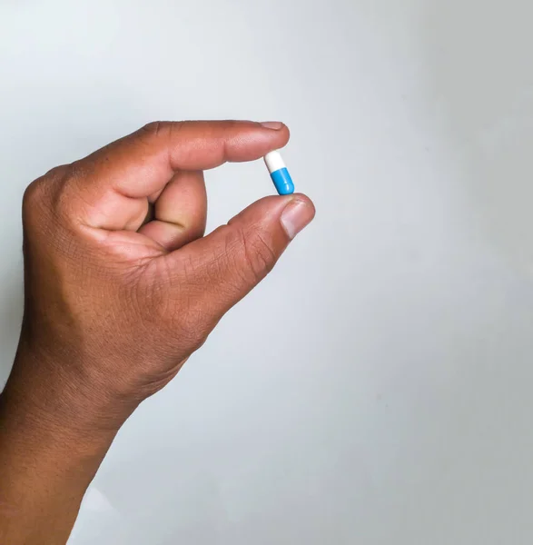 Close up on African male left hand holding a medicine capsule that\'s blue and white in color isolated on a white background. Hand holding pill capsule on white background