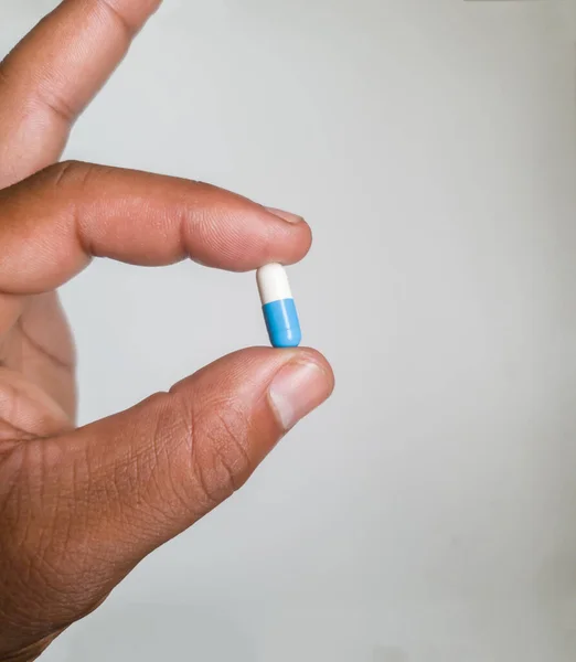Close up on African male left hand holding a medicine capsule that\'s blue and white in color isolated on a white background. Hand holding pill capsule on white background