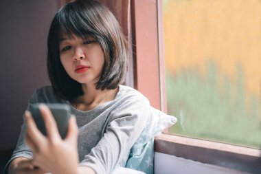 Yatak odasının penceresinde oturan bir kadın telefonuyla yüzünde kayıtsız bir ifade var..