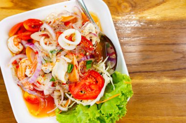 Tahta bir masaya yerleştirilmiş beyaz tabakta deniz ürünleri salatası, üst manzara, baharatlı Tayland yemeği..