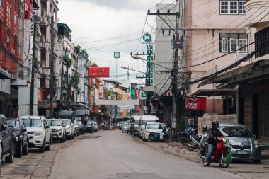 Maesot, Tayland - 3 Temmuz 2024: Yol kenarına park etmiş araçlar, geçen motosikletliler ve bulutlu bir gökyüzünün altında caddeyi kaplayan binalar.