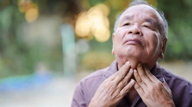 Yaşlı bir adam boğazına dokunuyor, muhtemelen rahatsızlık ya da endişeye işaret ediyor. Arka plan hafifçe bulanık, sakin bir atmosfer yaratıyor.