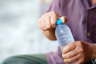 Plastik su şişesini açan kişi, tutucu ellerini gösteriyor. Ellere ve şişeye odaklanmış, geçmişi bulanık. Sahne ferahlatıcı duyguyu iletir.