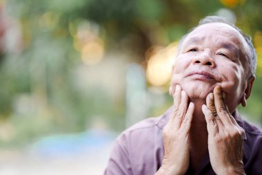 Düşünceli yaşlı bir adam yukarı bakarken nazikçe yüzüne dokunur, bir yansıma ve sükunet hissi verir..