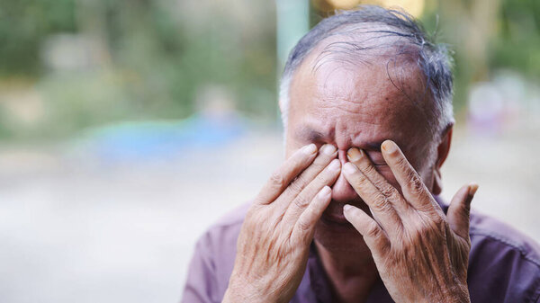 Пожилой человек закрывает лицо руками, передавая глубокие эмоции и созерцание в размытой наружной обстановке..